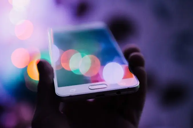 Zbliżenie na smartfon trzymany w prawej dłoni, ekran odbija rozmyte kolorowe światła w odcieniach fioletu, różu, zieleni i pomarańczu, w tle ciemne niewyraźne tło; Agencja marketingowa odnosi obrazek do usługi prowadzenie social media.